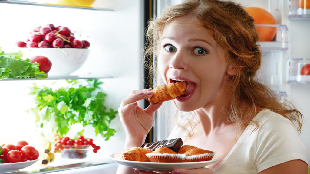 Женщина на фоне открытого холодильника ест круассан и держит в руке тарелку с кексами