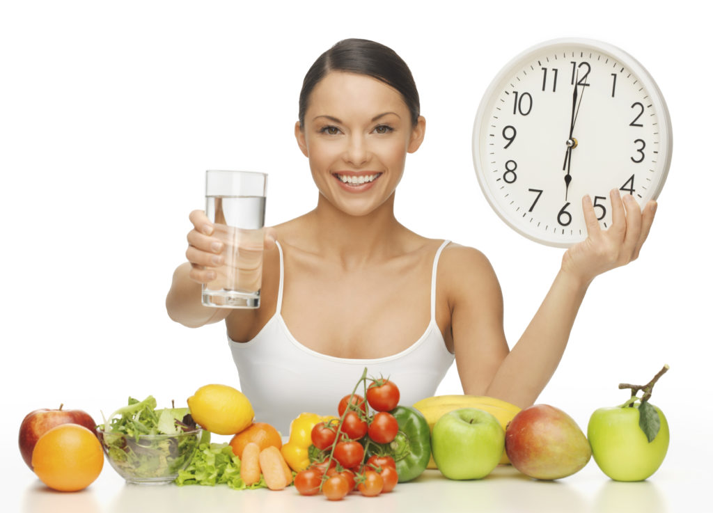 Девушка сидит за столом с овощами и фруктами и держит в руках стакан воды и часы