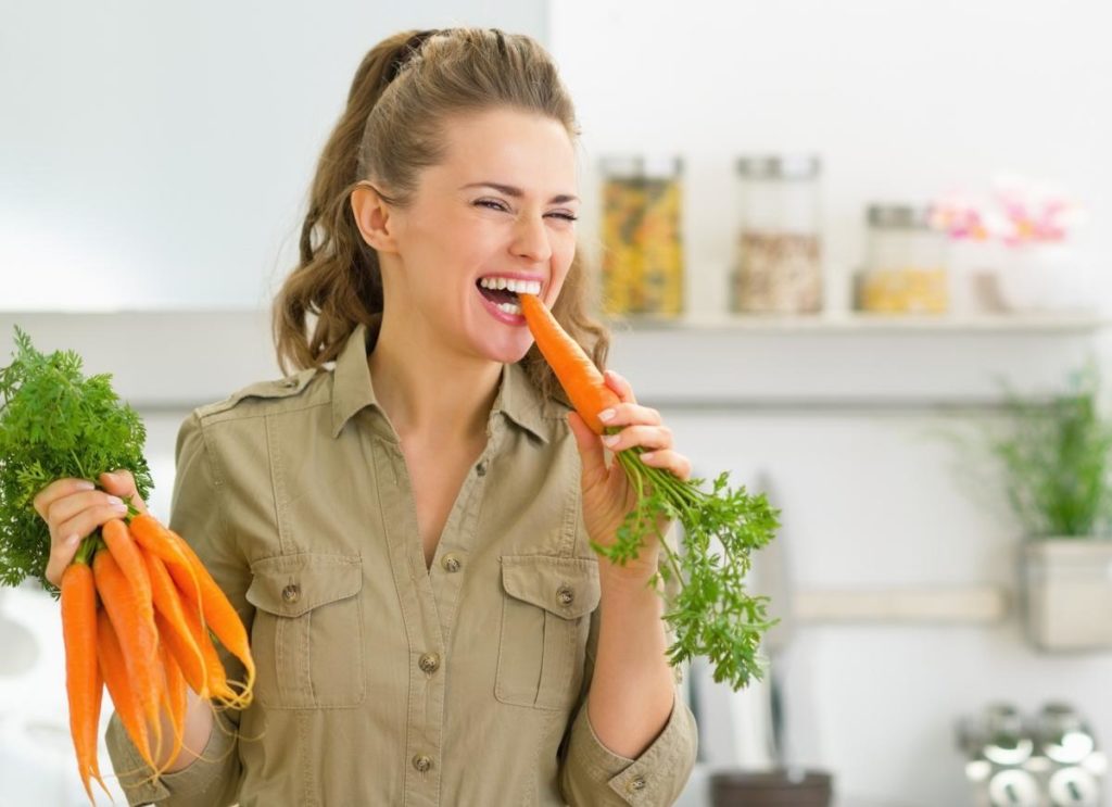 Девушка ест морковь на кухнке и держит в руке связку моркови