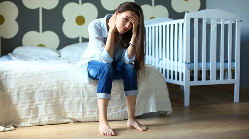 Уставшая молодая женщина сидит возле детской кроватки